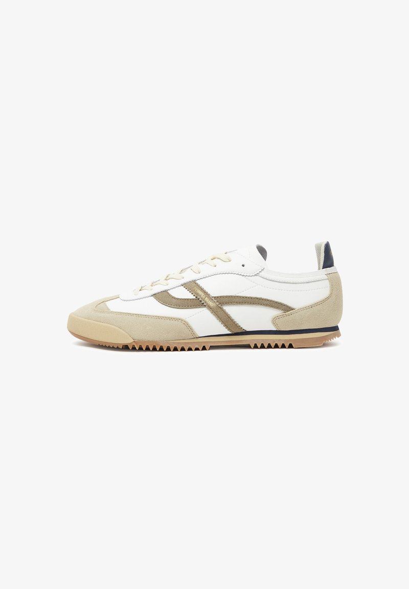 White sneaker with beige suede accents, featuring a textured rubber sole, minimal design, and contrasting brown side stripes.