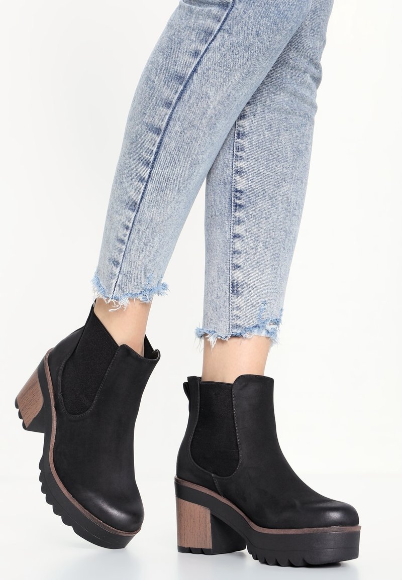 Bottines noires à talon épais en bois, avec des panneaux latéraux élastiques et une semelle texturée, portées avec un jean bleu clair à ourlet effiloché.