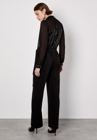 Schwarzer Jumpsuit mit durchsichtigen langen Ärmeln, mit Pailletten verziertem Einsatz im Rücken, taillierter Passform und geradem Bein. Hochhackige Schuhe sind im Lieferumfang enthalten.