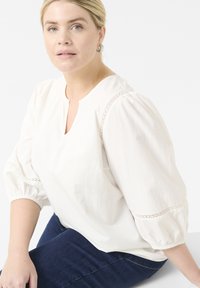 Valkoinen paita, jossa on V-pääntie, lyhyet puffihihat ja pitsiyksityiskohdat hartioilla. Yhdistetty tummansinisiin farkkuihin. Sujuva kankaan rakenne.