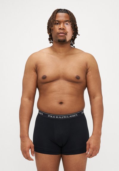 Boxer shorts neri in cotone con una fascia con logo. Modello con tono di pelle mediamente scuro si trova di fronte a uno sfondo bianco semplice, mostrando il prodotto.