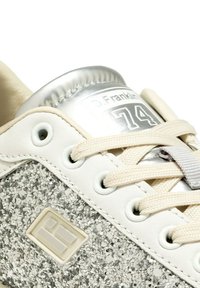 Sneaker bianca con accenti argentati, pannello laterale glitterato texturizzato, lacci beige e collare argentato imbottito. Presenta il logo e il numero 74.