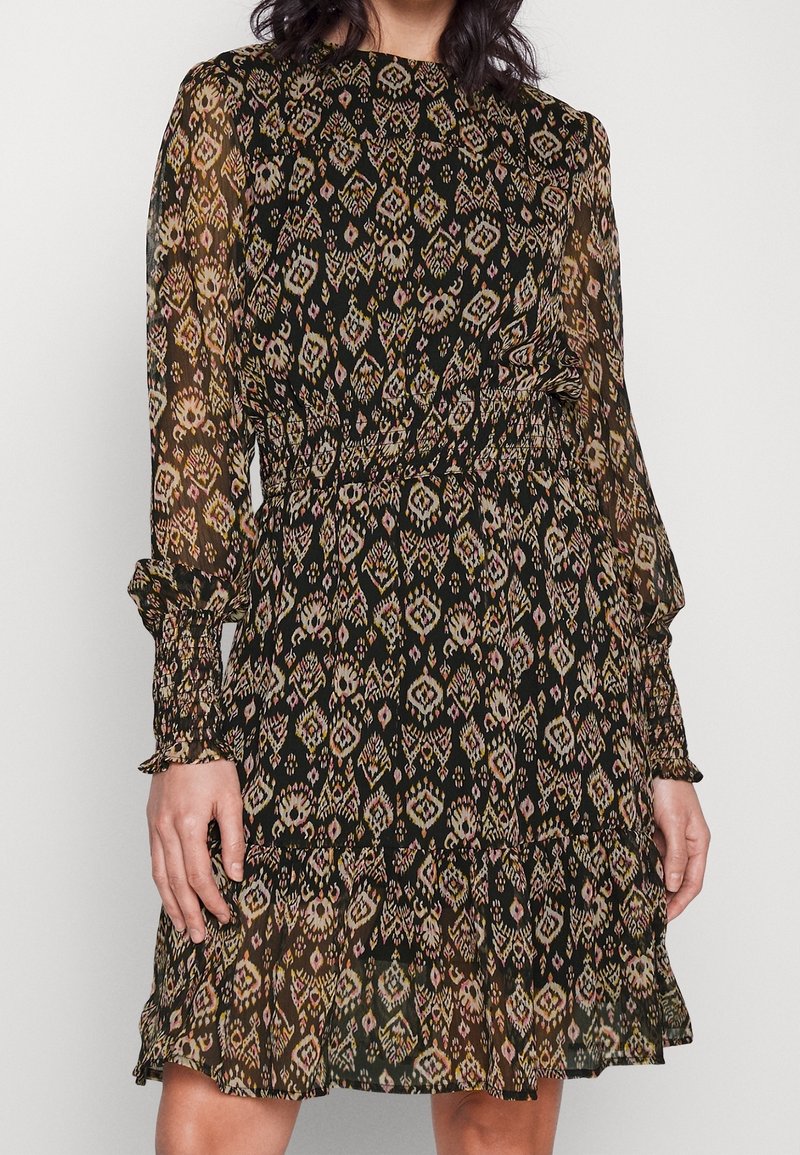 Femme portant une robe noire transparente à manches longues avec une taille froncée et un motif géométrique multicolore, debout devant un fond clair uni.