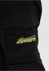 Black cargo pants with prominent yellow "Rocawear" embroidery on the side pocket. Features multiple pockets and a durable fabric texture.