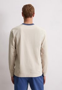 Uomo con capelli corti castano chiaro, che indossa una felpa crema e pantaloni blu, in piedi di fronte a una parete leggera e uniforme.