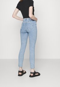 Žena v světle modrých skinny džínách s vysokým pasem, černém krátkém topu a černých sandálech na otevřenou špičku, stojící před jednobarevným pozadím.