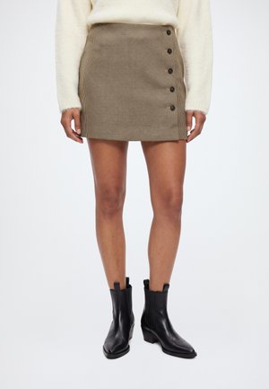 Modèle portant une minijupe marron à carreaux boutonnée sur le devant, un pull couleur crème et des bottines noires sur fond blanc.
