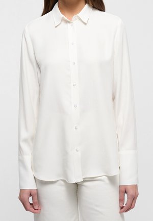 Vrouw draagt een wit overhemd met lange mouwen, knopen en een kraag, gecombineerd met een witte broek, staand tegen een effen lichte achtergrond.