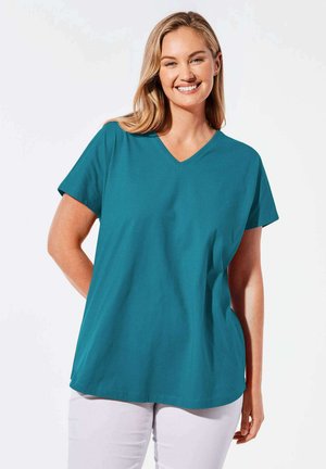 T-shirt teal à col en V et manches courtes, en tissu lisse. Coupe ample avec un ourlet droit, porté avec un pantalon blanc.