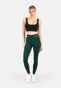 Zwart crop top en donkergroene leggings met hoge taille en "SMIL" logo op de tailleband. Model draagt witte sportschoenen, staand op een neutrale achtergrond.