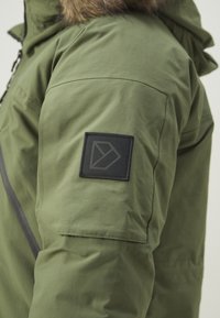 Parka verde oliva con superficie testurizzata, caratterizzata da una toppa quadrata nera sulla manica con un logo geometrico e cappuccio foderato di pelliccia.