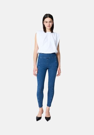 Donna in piedi che indossa un top bianco senza maniche con spalle imbottite, jeans skinny blu a vita alta e tacchi neri a punta.