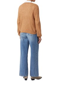 Eine Person trägt einen braunen Strickpullover, hellblaue weit geschnittene Jeans und beigefarbene, Absatz-Ankle-Boots. Sie steht mit dem Rücken zu einem weißen Hintergrund.