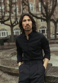 Cămașă neagră cu nasturi și guler, asortată cu pantaloni închiși la culoare. Materialul este neted, cu croială ajustată. Fundalul include copaci și clădiri.