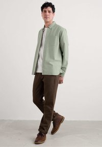 Lichtgroene knoop-down shirt met een borstzak, gelaagd over een wit shirt, gecombineerd met donkerbruine broek en bruine schoenen.