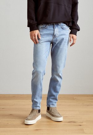 Hombre con vaqueros ajustados azul claro, botas beige de ante sin cordones y una sudadera negra con capucha, de pie sobre un suelo de madera frente a una pared lisa.
