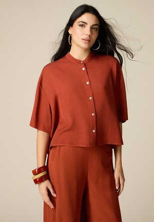 Donna con capelli lunghi e scuri indossa una camicia ampia color ruggine con bottoni e pantaloni abbinati, accessoriate con grandi orecchini a cerchio e braccialetti spessi.