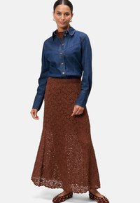 Chemise en denim à boutons sur le devant et col classique, associée à une jupe maxi en dentelle marron et des chaussures à enfiler avec des accents dorés.