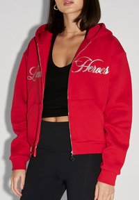 Rote Zip-Hoodie aus Baumwolle mit Kapuze, mit silberner Stickerei "Lucas" und "Heroes", sowie Fronttaschen. Schwarzes Oberteil darunter.