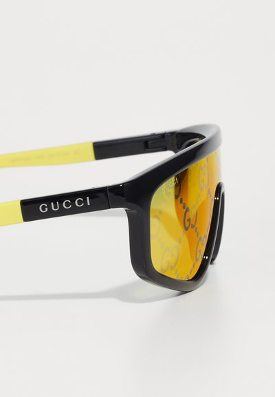 Gucci prillid musta raami ja kollaste kätega, millel on kõverad servad ja peegeldavad kollased objektiivid, millel on G logo muster.