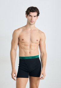 Schwarze Boxer-Brief mit einem grünen Calvin Klein Bund. Hergestellt aus weichem Stoff, haben sie eine glatte Textur und eine Länge bis zur Mitte des Oberschenkels.