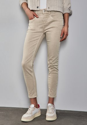 Persoon die strakke beige broek draagt, witte sneakers met roze accenten en een lichtbeige jas, staand tegen een effen achtergrond.