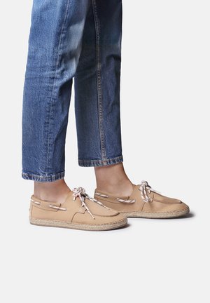 Personne portant un jean bleu court et des chaussures à enfiler couleur fauve avec des lacets blancs, debout sur un fond gris clair.