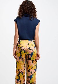Femme avec des cheveux frisés portant un haut sans manches bleu marine et un pantalon large taille haute à motifs floraux jaunes et roses, vue de dos.