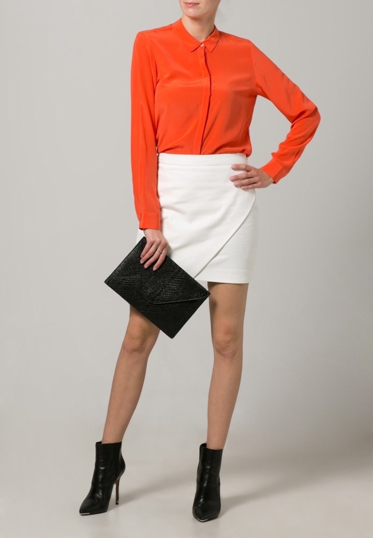 Blouse orange à manches longues associée à une jupe portefeuille blanche. Une pochette noire texturée et des bottines complètent la tenue.