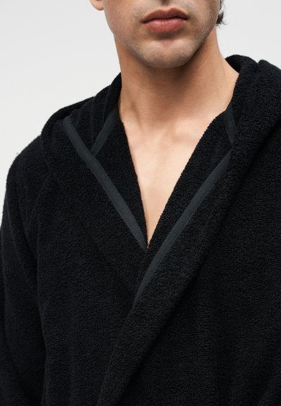 Bărbat tânăr purtând un halat negru cu glugă, dintr-un material texturat, vizibil de la gât până la partea superioară a pieptului, pe un fundal deschis.