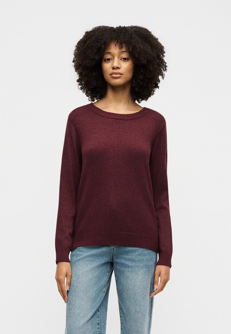 Femme aux cheveux bouclés portant un pull bordeaux à manches longues et un jean bleu clair, debout devant un fond uni gris clair.