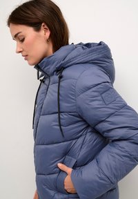 Puffer jacket in light blue, featuring a hood, black drawstrings, quilted texture, side pockets, and a logo patch on the sleeve.