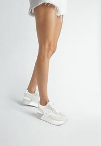 Sneakers atletici bianchi con tomaia testurizzata, dettagli in pelle scamosciata e una suola spessa e curvata per maggiore altezza e supporto.