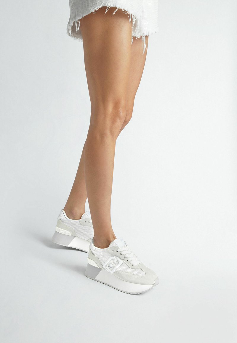 Sneakers atletici bianchi con tomaia testurizzata, dettagli in pelle scamosciata e una suola spessa e curvata per maggiore altezza e supporto.