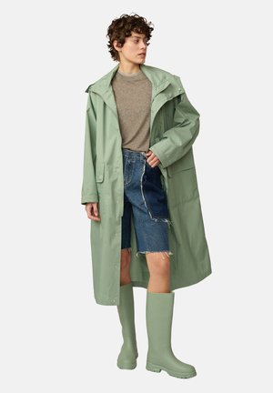 Un imperméable vert clair avec une capuche, associé à un haut gris, un short en denim et des bottes en caoutchouc vertes. Il présente des fermetures à pression et des poches.