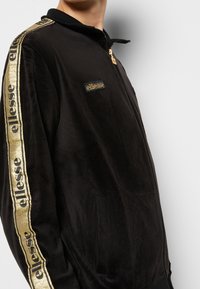 Svart velour zip-up jacka med guldfärgade accentstripes och logotyp på bröstet, med stående krage och sidofickor.