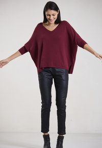 Burgunderfarbener übergroßer Strickpullover mit V-Ausschnitt und Dreiviertelärmeln, kombiniert mit schwarzen Lederhosen und Stiefeletten. Glatte Textur.