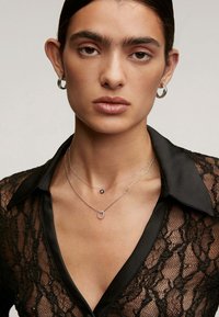 Una persona joven con cabello oscuro lleva pendientes de aro de plata, dos collares de plata en capas y una blusa de encaje negro con cuello de satén.