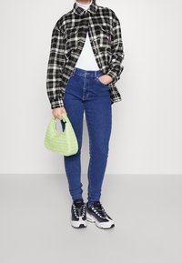Black and white plaid oversized shirt, white top, high-waisted blue jeans, green woven handbag, and black athletic shoes with blue accents.