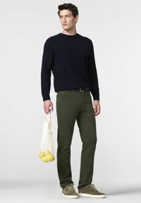 Un pull tricoté bleu marine, un pantalon ajusté vert olive et des baskets en suède gris. Tient un sac en filet blanc avec des objets jaunes. Design minimaliste.