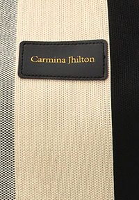 Correa de tela beige y negra con una superficie texturizada; presenta una etiqueta rectangular negra con texto en dorado grabado, "Carmina Jhilton."