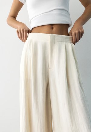 Egy személy lazán illeszkedő, magas derekú, krémszínű nadrágot igazít, fehér, rövid felsőt viselve, egy sima világos háttér előtt.