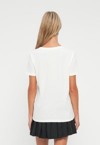 Γυναίκα με μακριά ξανθά μαλλιά, ντυμένη με ένα απλό λευκό κοντομάνικο πουκάμισο και μια μαύρη πλισέ φούστα, στέκ εται στραμμένη προς τα πίσω μπροστά από άσπρο φόντο.