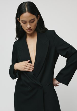 Blazer noir sur mesure avec un décolleté en V profond, des épaules structurées et des manches longues. Présente une fermeture à bouton et une texture de tissu lisse.