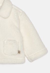 Biała, pluszowa kurtka z futra sztucznego z okrągłym kołnierzem. Posiada kwadratową kieszeń naszywaną i małą metkę logo blisko dołu.