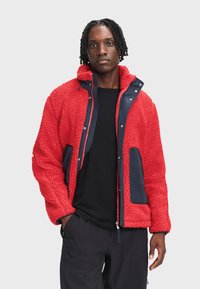 Czerwona kurtka polarowa o teksturowanej powierzchni, z granatowymi akcentami, dwoma przednimi kieszeniami, zapięciem na zamek i kapturem. Pod spodem czarna koszulka.