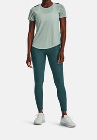 Ljust grönt luftigt kortärmat plagg med texturerad tyg och axelutskärningar, parat med mörk teal leggings med ett subtilt mönster och ankellängd.