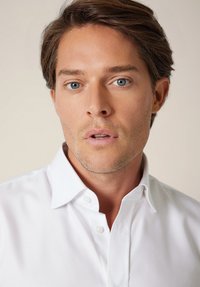 Biała koszula na guziki z klasycznym kołnierzykiem, gładka w fakturze i o prostym wzorze. Skupienie na nienagannym wyglądzie koszuli i schludnym kroju.