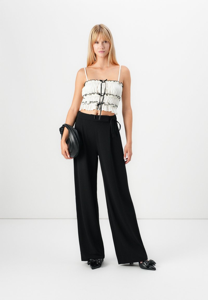 Witte cropped top met zwarte ruffle trim en een knoop aan de voorkant, gecombineerd met hoge zwarte wijde pantalon en zwarte puntige schoenen.