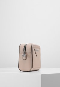 Vaaleanbeige suorakulmainen olkakassi mustalla reunuksella, jossa etutasku ja hopeinen vetoketju, esillä valkoisella jalustalla yksinkertaista taustaa vasten.